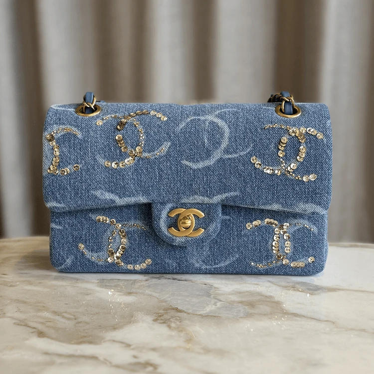Light blue denim Chanel Classic Flap handbag with gold sequin CC logo pattern, antique gold CC turn-lock clasp, and chain shoulder strap, displayed on a marble surface.